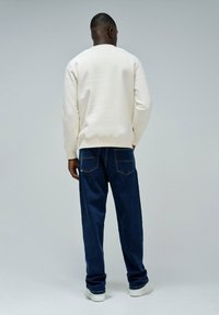 Man in een wit sweatshirt, donkerblauwe jeans en witte sneakers staat met zijn rug naar de camera tegen een effen grijze achtergrond.