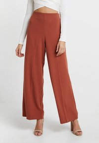 Femme portant un pantalon large taille haute couleur rouille avec des talons ouverts beiges et un haut court à manches longues blanc, se tenant devant un fond blanc.