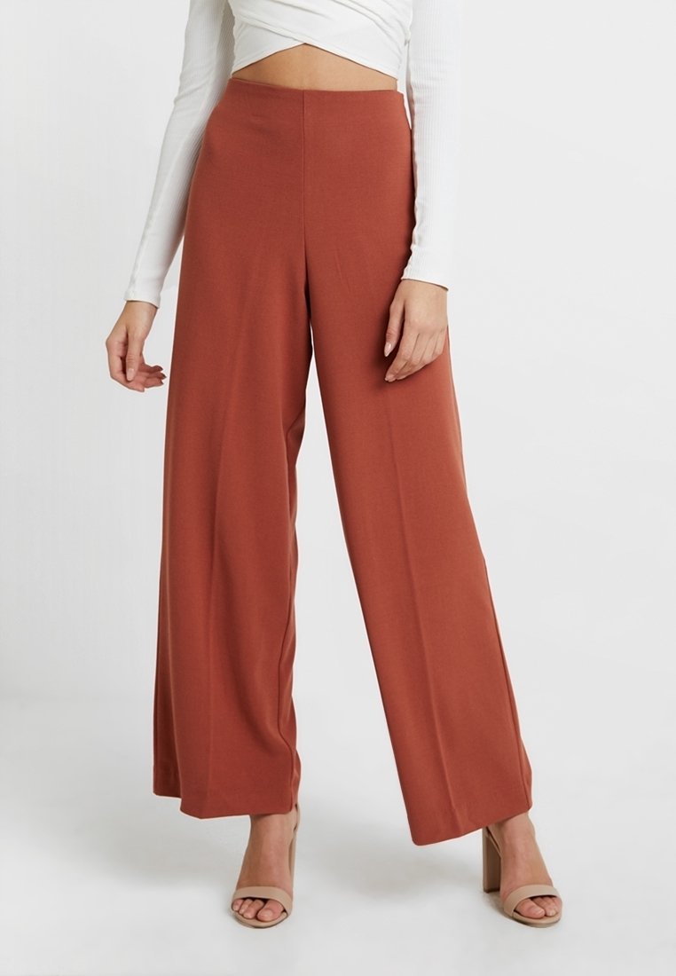 Femme portant un pantalon large taille haute couleur rouille avec des talons ouverts beiges et un haut court à manches longues blanc, se tenant devant un fond blanc.