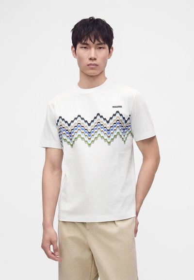 Νέος άνδρας που φοράει λευκό T-shirt με μπλε, μαύρες και πράσινες ζιγκ ζαγκ ρίγες και μπεζ παντελόνι σε απλό λευκό φόντο.