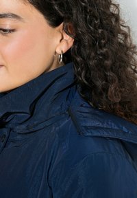 Laivastonsininen takki, jossa on korkea kaulus, kiiltävä tekstuuri, vetoketjusomisteet ja vettä hylkivä viimeistely; yhdistetty renkaankorvakoruihin.