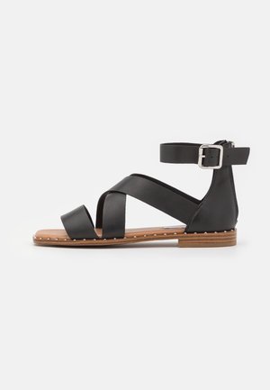 Black leather sandal with cross straps, ankle buckle, and flat sole. Features a wooden base with metal studs along the edge.