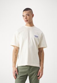 Cream-colored cotton T-shirt with short sleeves, featuring a blue "Originals" logo on the left chest. Relaxed fit and standard crew neckline.