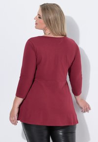 Bordeauxfarbene Langarmbluse mit Rundhalsausschnitt und A-Linie-Silhouette, aus weichem Stoff gefertigt. Kombiniert mit schwarzen, eng anliegenden Hosen.