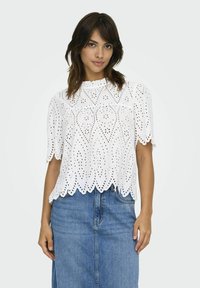 Weiße Bluse aus Eyelet mit gewelltem Saum und kurzen Ärmeln, mit dekorativen Ausschnitten und einem Rüschen-Halsausschnitt, kombiniert mit einem Jeansrock.