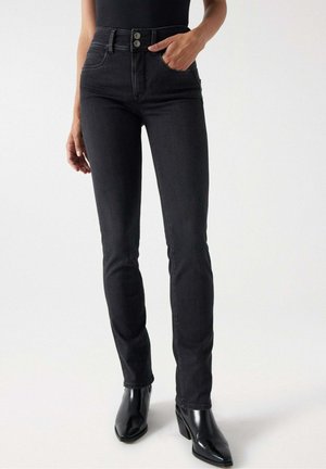 Personne portant un jean skinny noir taille haute avec fermeture à double bouton, associé à des bottines noires à bout pointu.