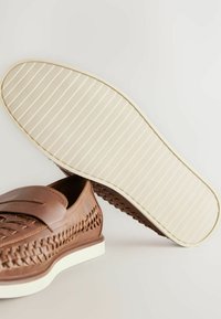 Zapato de cuero marrón con textura trenzada, suela blanca y diseño de cordones. Presenta una lengüeta acolchada y un detalle estriado en la suela.
