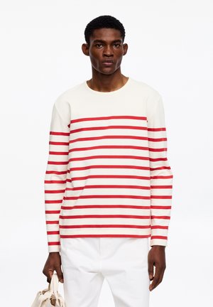 Man staat met een beige canvas draagtas, draagt een wit shirt met lange mouwen en rode horizontale strepen en witte broek tegen een witte achtergrond.