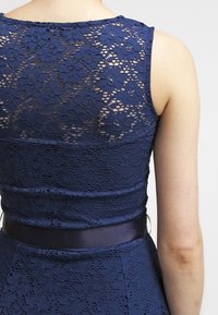 Robe en dentelle bleu marine avec un motif floral, une taille cintrée et une ceinture en satin. Le dos présente un design de dentelle superposée avec une texture douce.