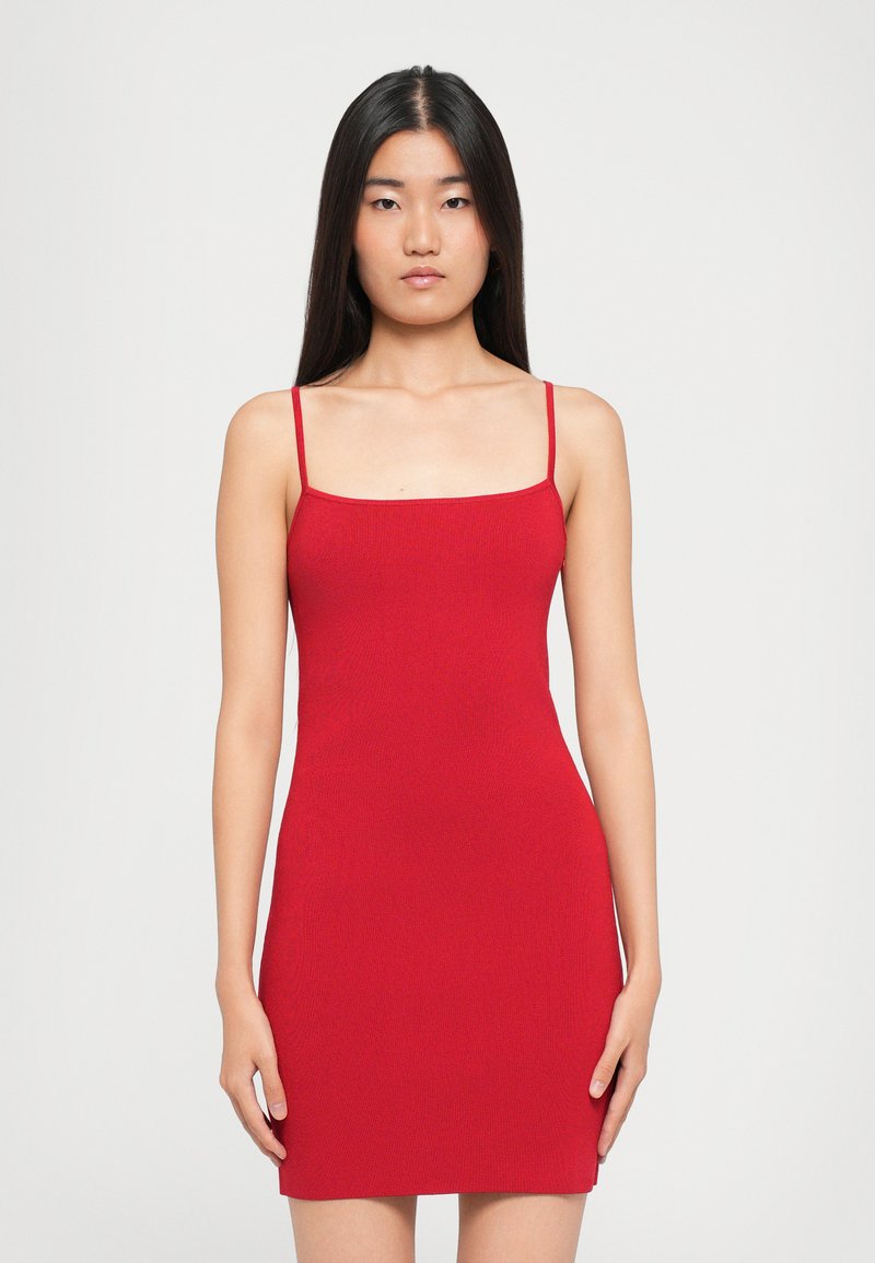 Donna con lunghi capelli neri che indossa un vestito rosso aderente senza maniche con spalline sottili, in piedi di fronte a uno sfondo bianco semplice.