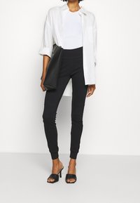 Černé legíny s přiléhavým střihem, v kombinaci s bílým přiléhavým tričkem a nadměrnou bílou košilí na knoflíky, doplněné černými lodičkami na podpatku.