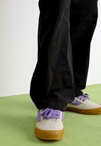 Adidași gri deschis cu șireturi și accente purpurii, având un superior texturat și o talpă din cauciuc de tip gumă, asortați cu pantaloni negri relaxați.