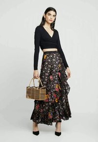 Mulher a usar uma blusa preta de manga longa e um maxi saia floral preta, segurando uma bolsa retangular de madeira com alças de bambu.