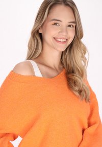 Pulover supradimensionat, cu umerii descoperiți, de culoare portocalie aprinsă, cu textură tricotată, având un decolteu larg și o curea albă vizibilă dedesubt.