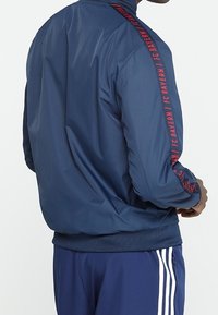 Navy sports jacket with red FC Bayern logo along the sleeves, featuring a smooth texture and ribbed cuffs, paired with matching pants.