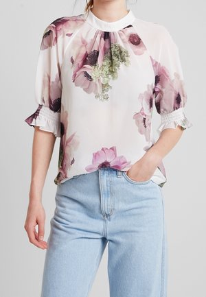 Femme portant un chemisier blanc avec un imprimé floral rose et vert et des manches bouffantes, associé à un jean bleu clair, main dans la poche.