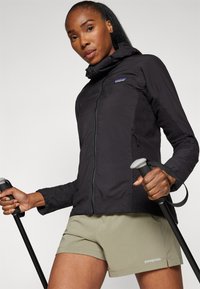 Patagonia lightweight black jacket with textured side panels, full zip, and hood. Pair of light green shorts and trekking poles visible.