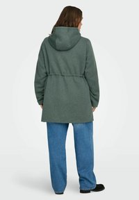 Zelena flis jakna s kapuco, zategnjena v pasu, z dolgimi rokavi in sproščenim krojem, v kombinaciji z modrimi kavbojkami in črno obutvijo.