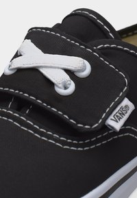 Κοντινό πλάνο μαύρου αθλητικού παπουτσιού Vans με λευκά σταυρωμένα κορδόνια, λευκά ραφτά και ύφασμα με λογότυπο στη πλευρά κοντά στη σόλα.