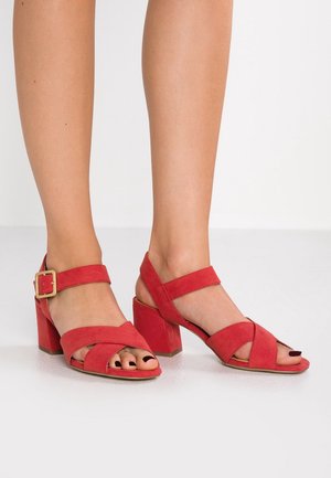Jambes portant des sandales à talons carrés en daim rouge avec brides à la cheville et boucle dorée, montrant des ongles de pieds vernis sur un sol blanc.