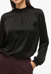 Schwarze Satinbluse mit gerafftem hohen Ausschnitt, langen Ärmeln mit elastischen Bündchen und lockerer Passform, kombiniert mit burgunderfarbenen Hosen.