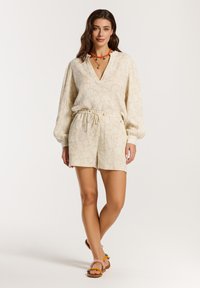 Completo corto beige chiaro con top a scollo a V a fantasia e pantaloni corti con cintura elastica; abbinato a sandali colorati e una collana multicolore.