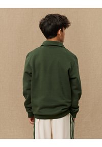 Mørkegrønn sweatshirt med krage, lange ermer og ribbestrikket kant. Bakfra vises glatt stofftextur og løst fit rundt midjen.