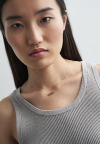 Kuldne kaelakee, millel on ristkülikukujuline ripats, kantud heledast hallist soonilisest topist, näidates sujuvat tekstuuri ja õrna kettide disaini.