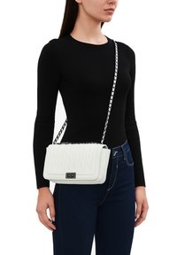Borsa a tracolla bianca trapuntata con catena metallica, di forma rettangolare, e una chiusura in metallo nera. Indossata sopra una maglia nera a maniche lunghe.