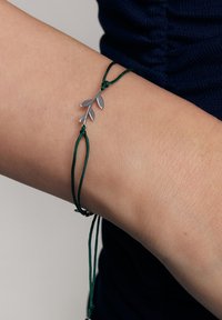 Srebrna bransoletka z zawieszką w kształcie liścia z zielonym regulowanym sznurkiem. Posiada delikatny wzór liścia i detale w formie węzłów. Powierzchnia jest gładka i metaliczna.