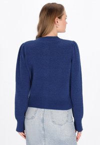 Maglione lavorato a maglia blu navy con maniche a sbuffo, colletto e orlo a costine, con una finitura testurizzata. Abbinato a una gonna di jeans chiara.