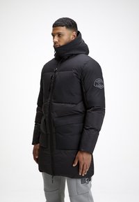Cappotto puffo nero con colletto alto e cappuccio regolabile, caratterizzato da una grande tasca frontale, chiusura con zip e patch-logo sulla manica.