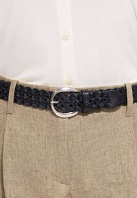 Marynarski pleciony pasek ze skóry z okrągłą srebrną klamrą, zestawiony z beżowymi teksturowanymi spodniami i białą koszulą na guziki.