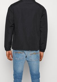 Czarna kurtka bomberka z elastycznymi mankietami i dołem, o gładkim wykończeniu. W zestawie z jasnoniebieskimi dżinsami z dekoracyjnym przeszyciem na kieszeniach.