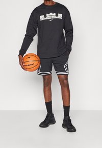T-shirt nera a maniche lunghe con motivo geometrico bianco sul petto, abbinata a pantaloni corti neri con bordini bianchi e scarpe da ginnastica nere, mentre tiene un pallone da basket.