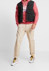 Svart väst med flera fickor över en röd huvtröja, beige byxor och vita skor med svarta och orange detaljer. Enfärgad bakgrund.