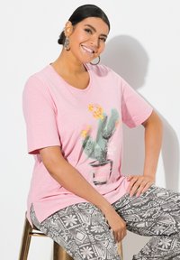 Camiseta de manga corta rosa con un gráfico de cactus con flores, combinada con pantalones grises con patrón. Tejido suave con corte relajado.