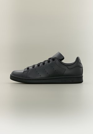 Μαύρο sneaker Adidas Stan Smith χαμηλού τύπου με κορδόνια, διάτρητες τρεις ρίγες, δερμάτινο επάνω μέρος και χαρακτηριστική ετικέτα στη φτέρνα και σόλα.