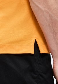 Κοντινή λήψη του χεριού ενός ατόμου που φοράει πορτοκαλί πουκάμισο με μικρή πλευρική σχισμή, συνδυασμένο με μαύρο παντελόνι.