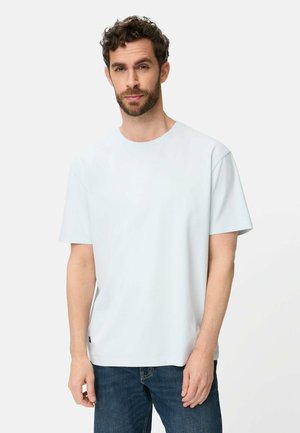 Mand med kort krøllet hår og skæg iført en enkel hvid T-shirt med korte ærmer og blå jeans, stående mod en hvid baggrund.