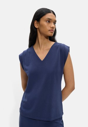 Kobieta z długimi ciemnymi włosami, ubrana w bezrękawnik w kolorze granatowym z dekoltem w serek, stojąca z rękami za plecami, spoglądająca na bok.