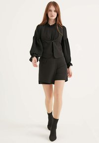 Schwarze Weste mit einem Reißverschluss und Taschen, kombiniert mit einem passenden schwarzen Rock. Das Outfit hat Puffärmel und knöchellange Socken.