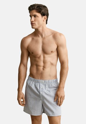 Oberkörperfreier Mann, der blaue und weiße vertikal gestreifte Boxershorts trägt, steht vor einem einfachen hellen Hintergrund und schaut zur Seite.