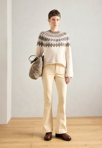 Πλέκτο πουλόβερ σε κρέμα με μπλε και καφέ γεωμετρικά σχέδια, συνδυασμένο με μπεζ καμπάνα παντελόνι και καφέ παντόφλες. Κρατάει μια μεγάλη γκρι τσάντα.