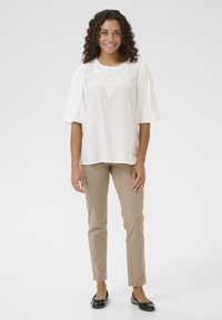Blusa bianca con dettagli in pizzo sul collo, maniche a sbuffo, abbinata a pantaloni fitted beige chiaro e scarpe ballerine nere.