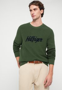 Zielony sweter dzianinowy z okrągłym dekoltem i granatowym logo "Hilfiger" z przodu, połączony z jasnobeżowymi spodniami i brązowym paskiem.