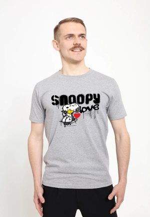 Mężczyzna w szarej koszulce z napisem "Snoopy love" i Snoopy trzymający czerwone serce, patrzy lekko w prawo i uśmiecha się.