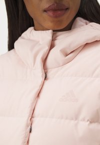 Svijetlo ružičasta jakna s punjenjem, visokog ovratnika, sa vidljivim kopčanjem na gumb i utisnutim Adidas logotipom u gornjem lijevom kutu.
