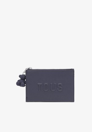 Cartera de piel texturizada azul marino con el logo "TOUS" en relieve, cierre de cremallera y un charm en forma de oso adjunto como decoración.