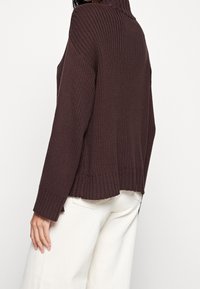Brauner, gerippter Strickpullover mit hohem Kragen, langen Ärmeln und einem leicht gekürzten Rücken. Mit seitlichen Schlitzen und einer strukturierten Oberfläche.